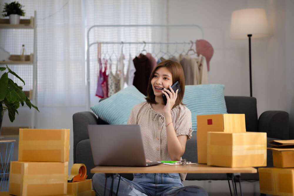 An online seller on her mobile phone while she's sitting at a table on which sits a laptop and some boxes