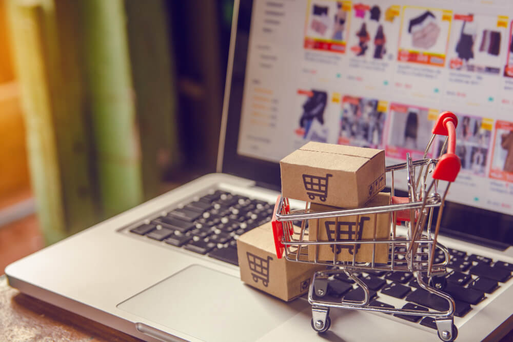 online-wholesale-stire-wholesale-pricing - AdTribes A laptop, its screen showing an online store, on which sits a miniature cart filled with tiny boxes